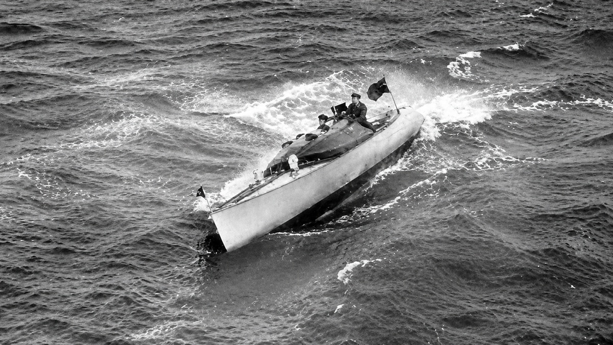 A small submarine is partially surfaced in rough ocean waters, with several crew members visible on top. Waves splash against the vessel as it moves through the sea.