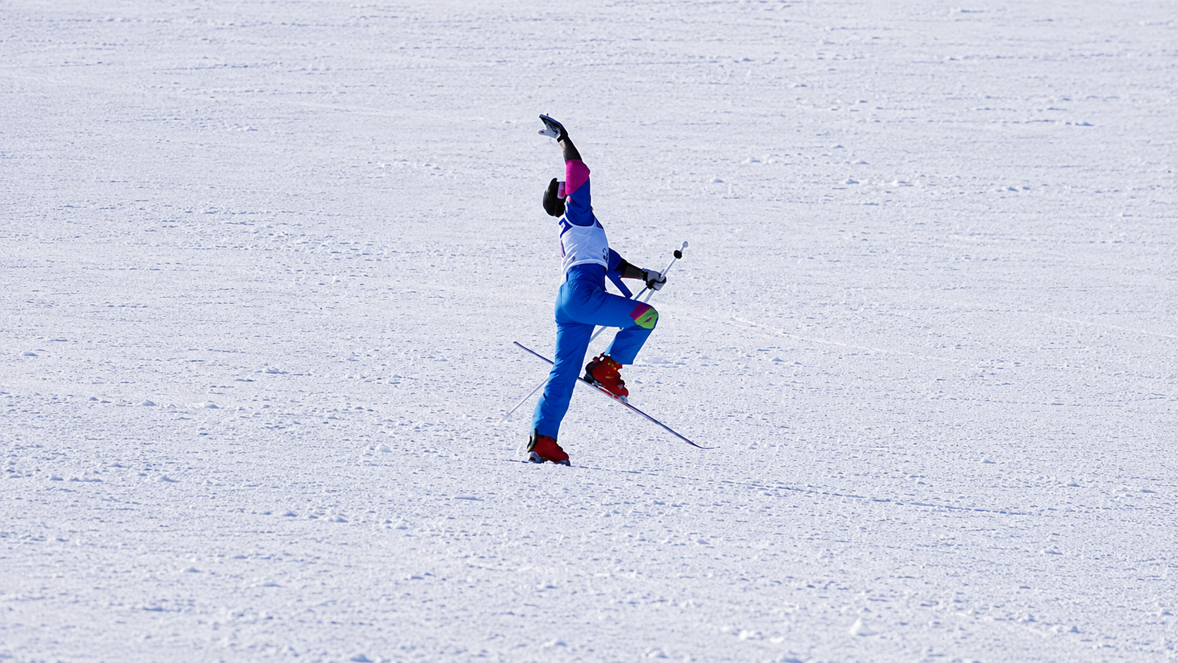 A skier in blue pants, a white top, and red boots is mid-fall on snowy terrain, with one arm raised and a ski pole extended, while a ski is off the ground.