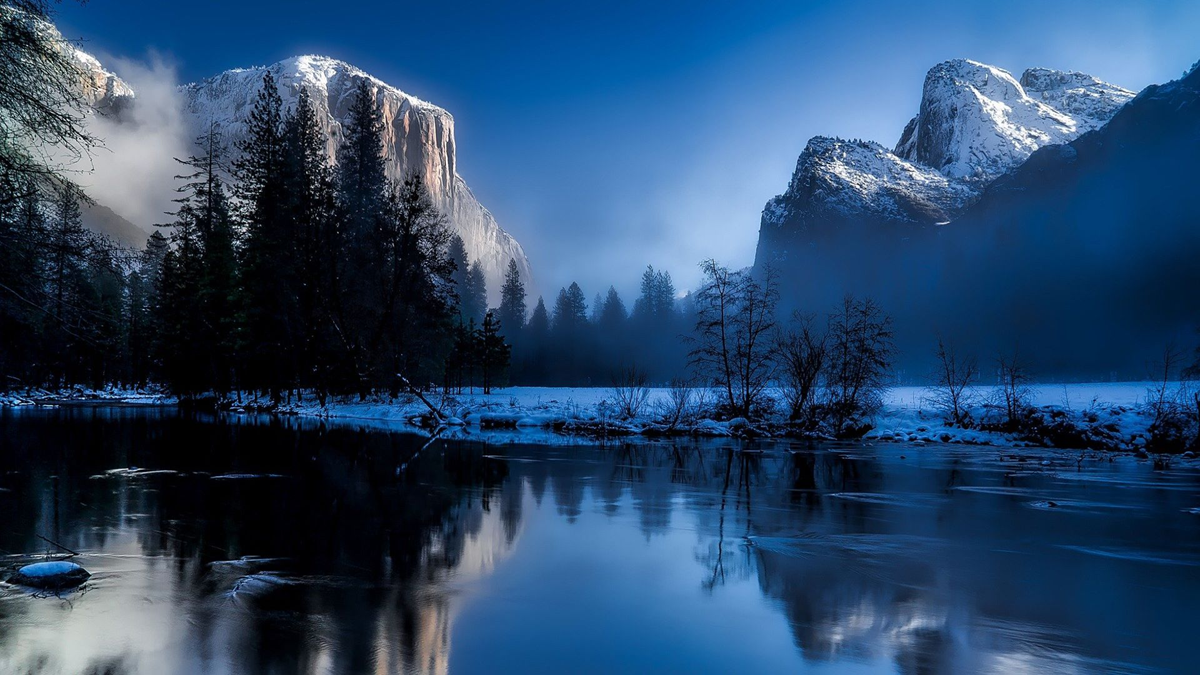 Snow-covered mountains rise above a tranquil river at dusk, reflecting pine trees and frosty banks. Mist hugs the landscape under a deep blue sky, creating a peaceful, wintery scene.