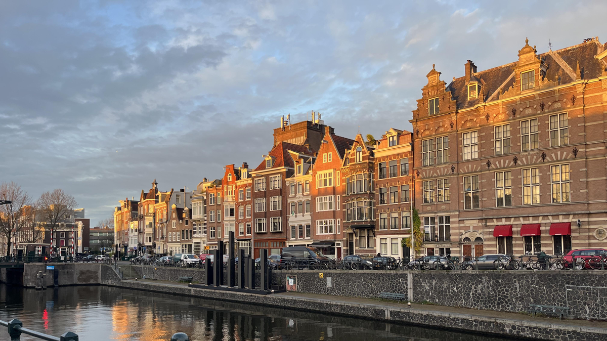 Rows of traditional Dutch buildings with red roofs line a canal in Amsterdam at sunset, reflecting warm light. Bicycles are parked along the water and clouds fill the sky.