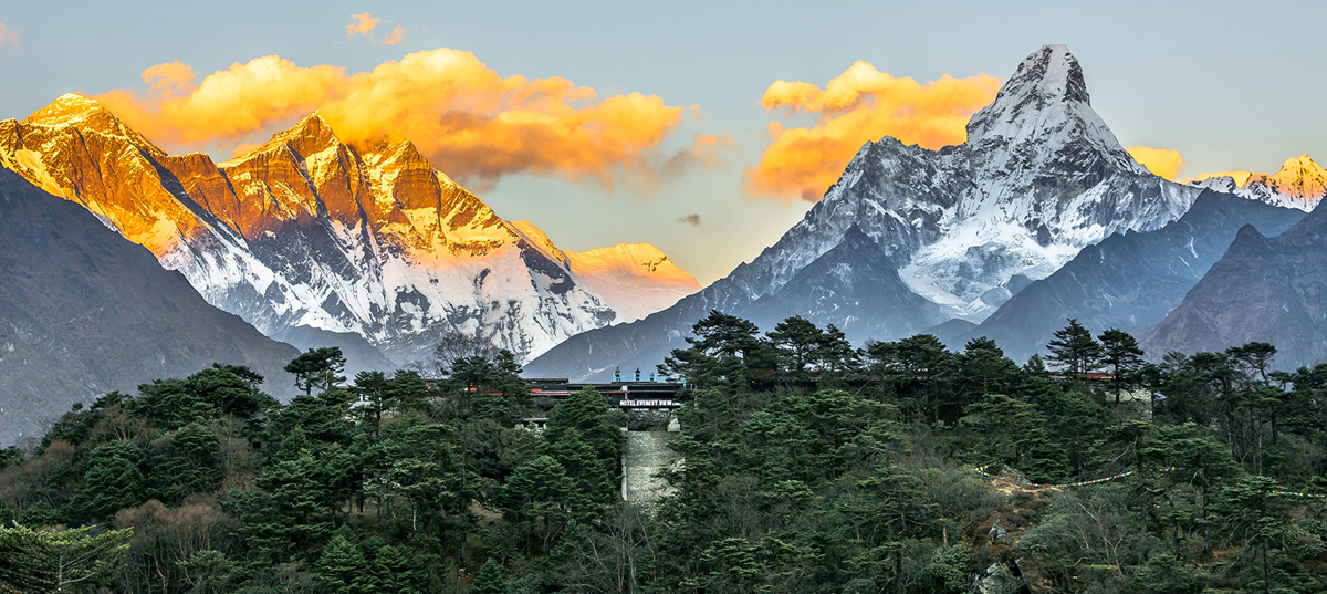 Snow-capped Himalayan mountains at sunrise, with golden light on peaks and a forested hill with buildings in the foreground. Orange clouds drift above the mountains.