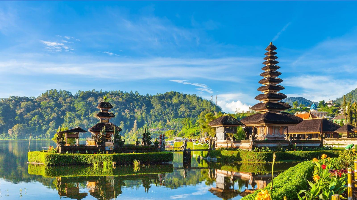 A lakeside Hindu temple with multi-tiered pagoda roofs is surrounded by lush greenery, mountains, and a clear blue sky, reflected in calm water.