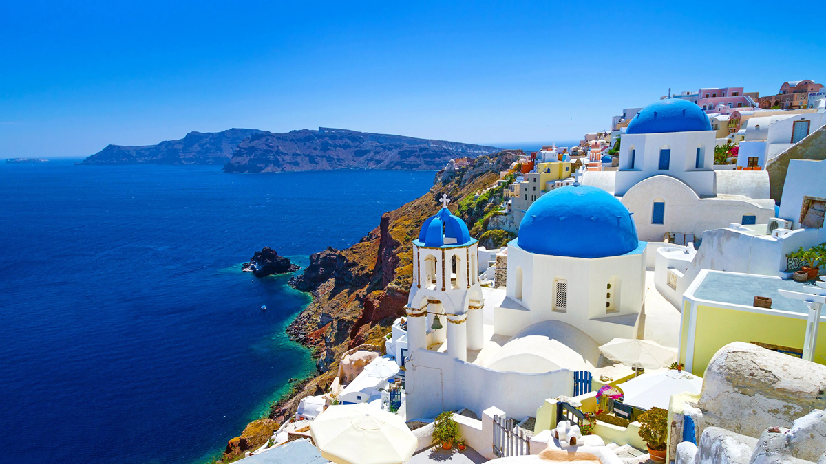White buildings with blue domes overlook a deep blue sea and rocky coastline in Santorini, Greece, under a bright, clear sky.