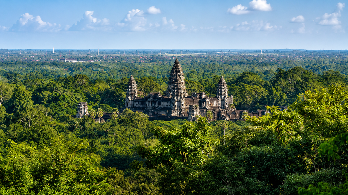 Ancient stone temple complex with tall towers rises above dense green jungle under a blue sky with scattered clouds, seen from a distance.