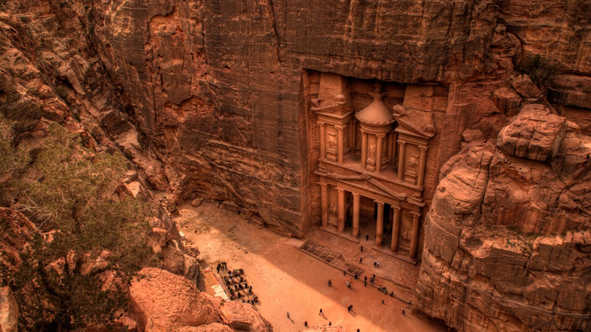 An aerial view of Petra’s ancient rock-cut Al-Khazneh temple in Jordan, surrounded by towering sandstone cliffs, with tourists gathered at its base.