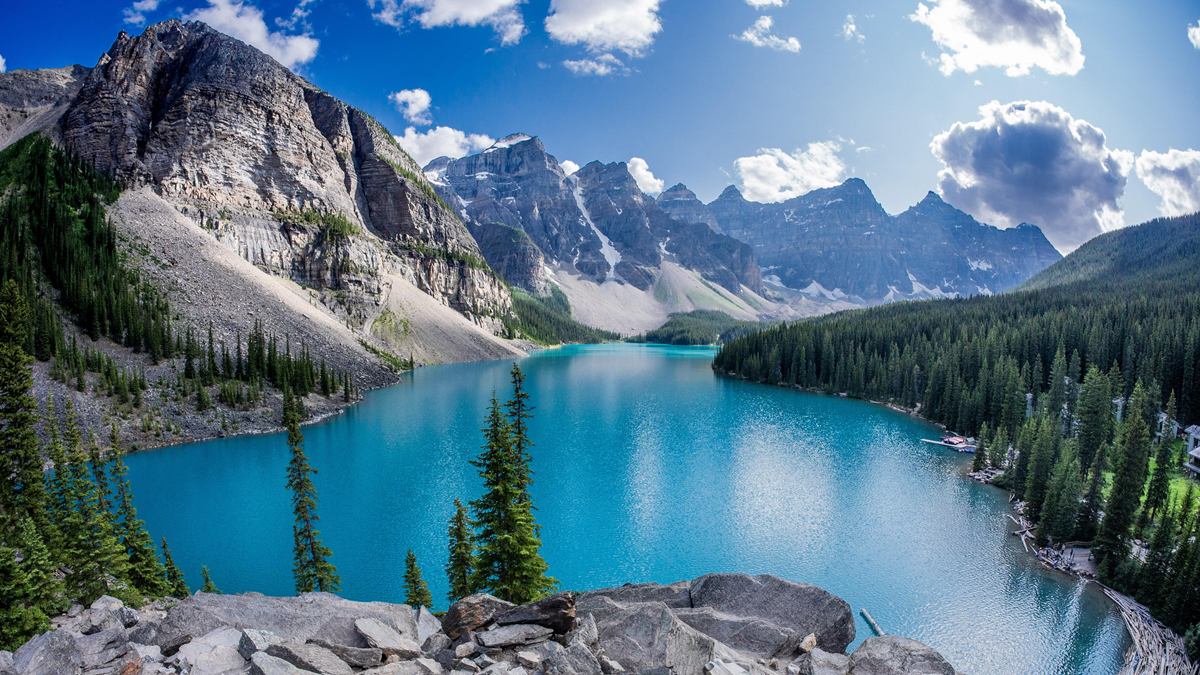 Clear blue lake surrounded by evergreen trees and tall rugged mountains under a bright partly cloudy sky. Rocky shoreline and forested hills frame the vivid scenery.