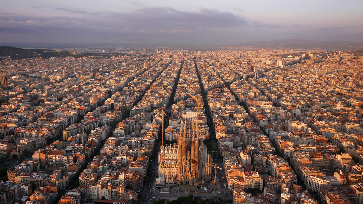 Aerial view of Barcelona at sunset, showing grid-like city streets and the Sagrada Familia basilica at the center, surrounded by densely packed buildings.