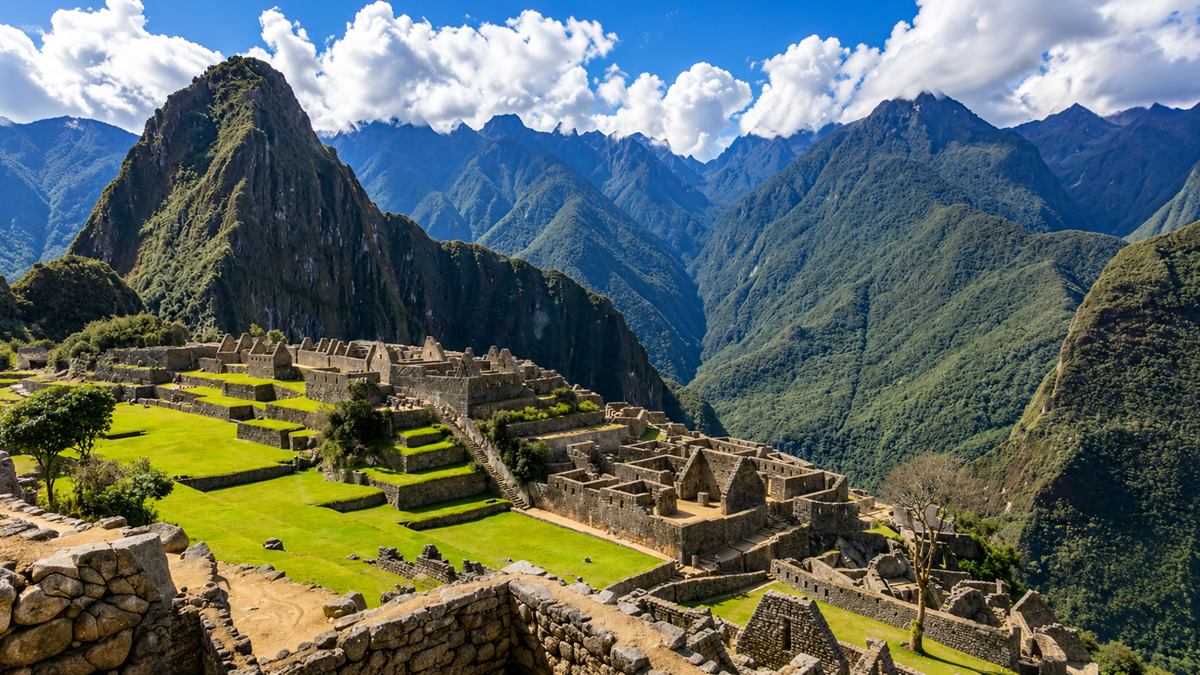 Ancient stone ruins of Machu Picchu sit atop a lush, green mountain ridge, surrounded by dramatic Andean peaks under a partly cloudy blue sky.