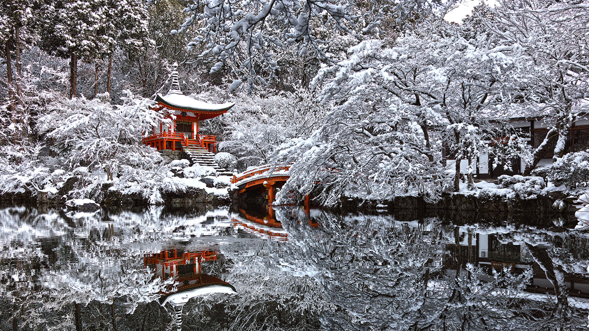 A red Japanese pagoda and arched bridge stand beside a pond, surrounded by snow-covered trees. Their reflection is visible in the still water, creating a serene winter landscape.