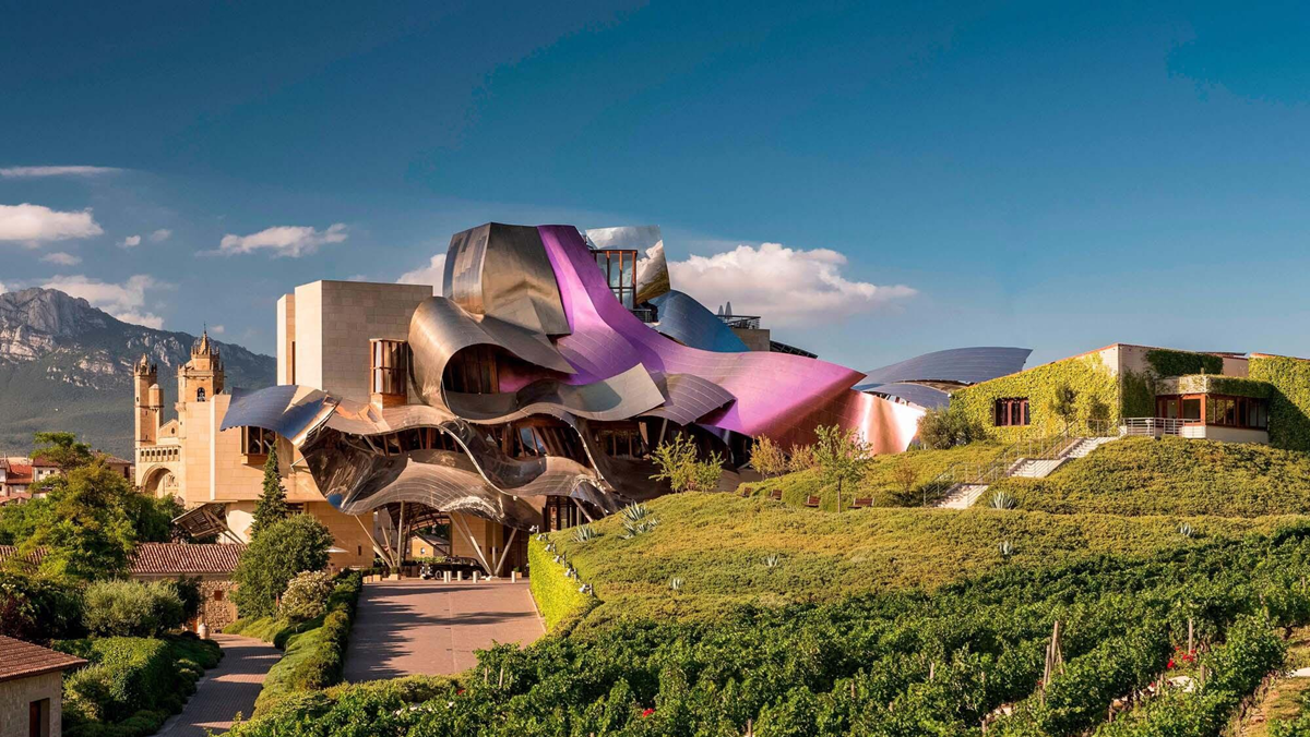 A modern building with flowing metallic surfaces in silver and purple sits amid green vineyards, with a historic stone tower and mountains in the background under a blue sky.