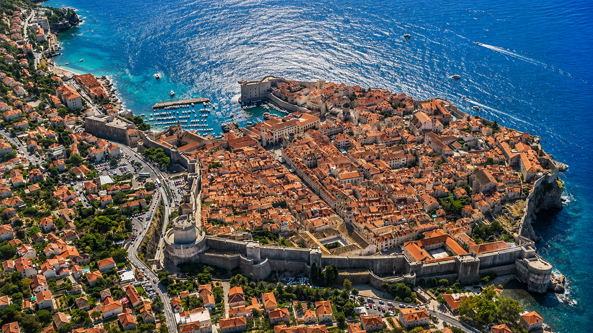 Places. Aerial view of a historic walled city with orange rooftops beside the blue Adriatic Sea, featuring harbors, stone buildings, and winding streets surrounded by lush green hills.