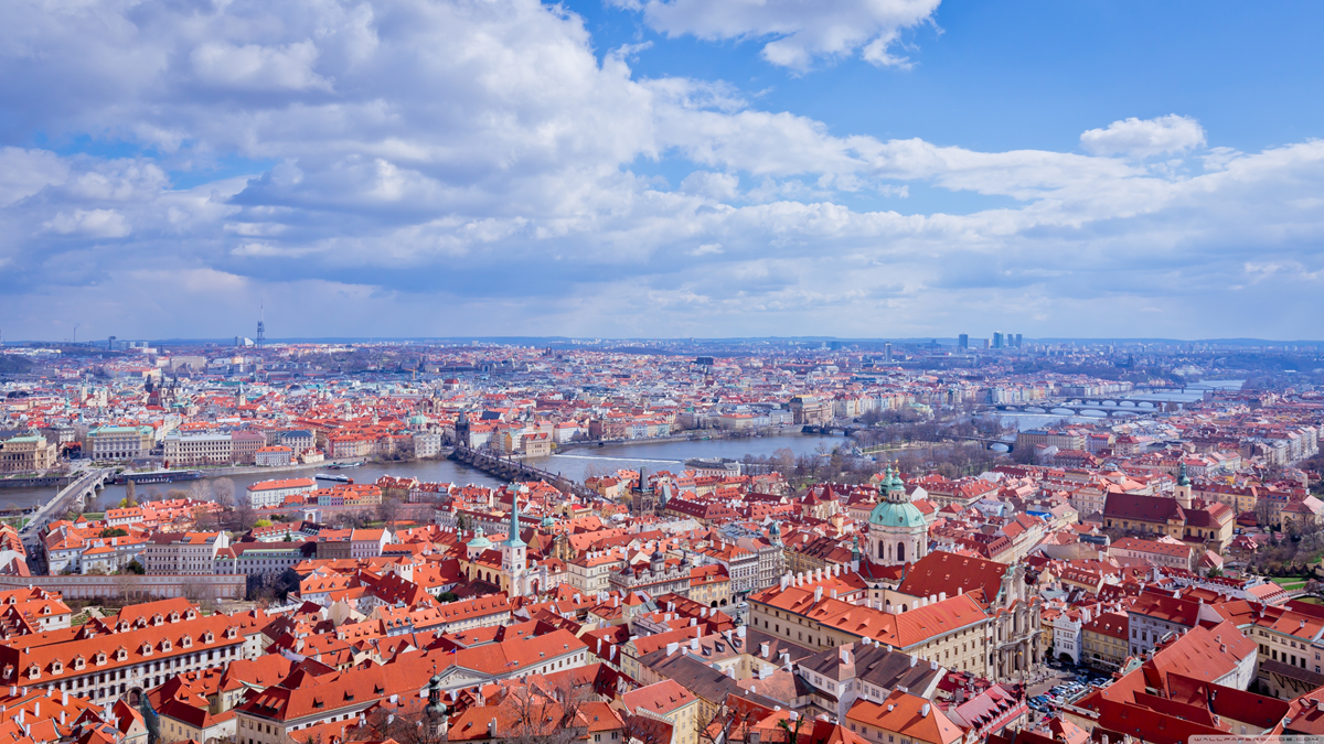 A panoramic view of Prague with red-roofed buildings, the Vltava River, historic bridges, and church domes under a partly cloudy sky.