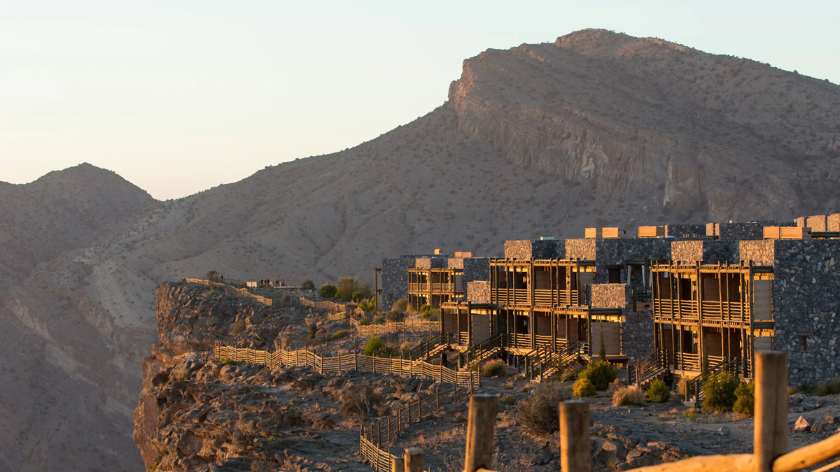 Modern stone buildings with wooden accents are perched on a rocky hillside, overlooking a rugged mountain landscape under soft sunlight.