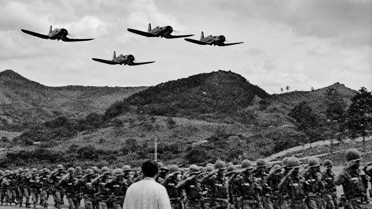 Soldiers in uniform salute as three military planes fly overhead against a backdrop of hills and cloudy sky. A person stands facing the group.