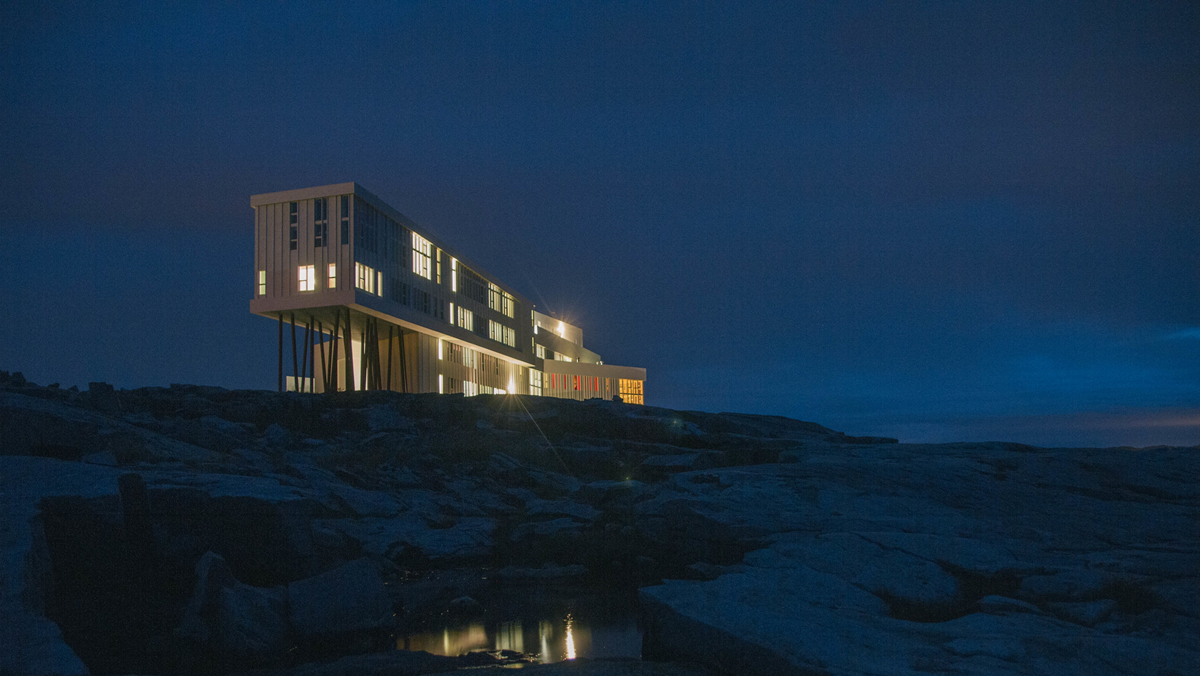 A modern, elevated building with illuminated windows stands on rocky terrain at night, set against a dark, cloudy sky.