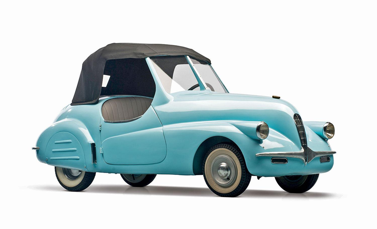 A vintage, light blue three-wheeled car with a black fabric convertible top, small tires, and a compact, rounded body is shown against a white background.