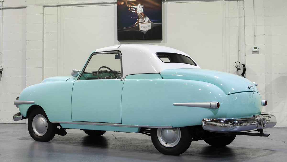 A vintage light blue and white two-door car is parked indoors on a gray floor, with white brick walls and a framed photo hanging above the car.