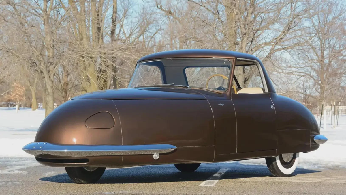 1940s- A vintage brown car with a rounded, streamlined design is parked outdoors on a snowy day, with bare trees in the background.