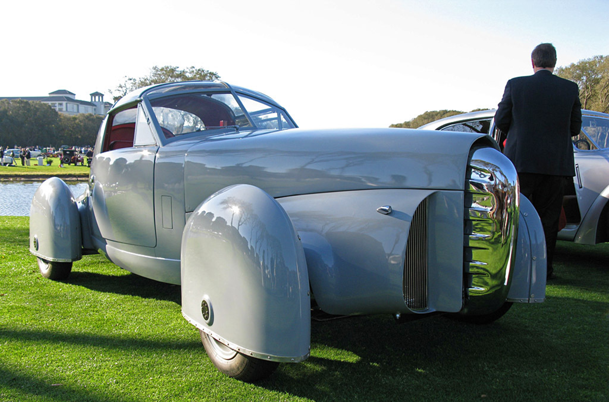 A vintage silver convertible car with large, rounded fenders and a chrome grille is displayed on grass at an outdoor event. A person stands nearby, facing away.