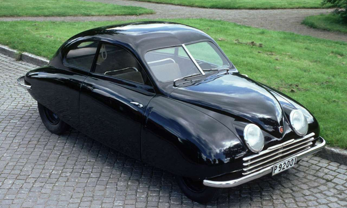 A vintage black Saab car with rounded body, large front fenders, and two prominent circular headlights parked on a cobblestone street next to a grass lawn.