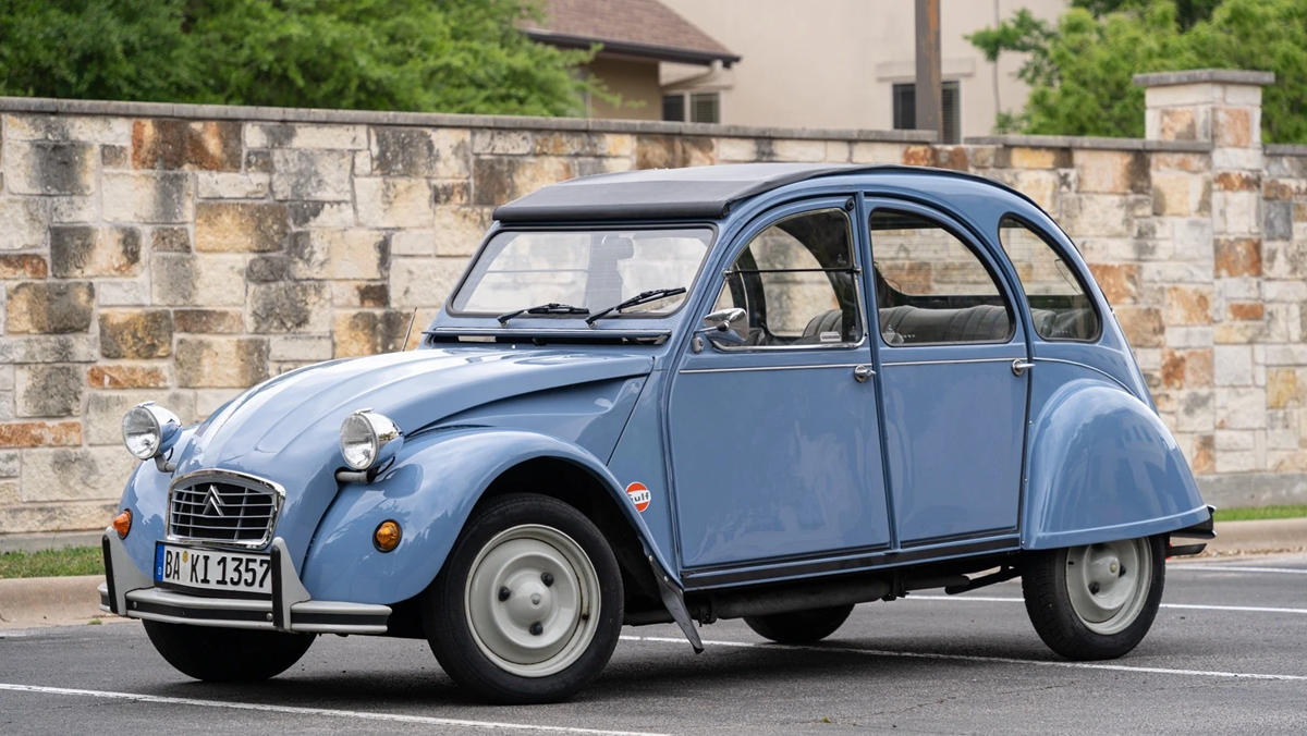 A light blue vintage Citroën 2CV car is parked in front of a stone wall and greenery. The car has a rounded body, exposed wheels, and a fabric roof.