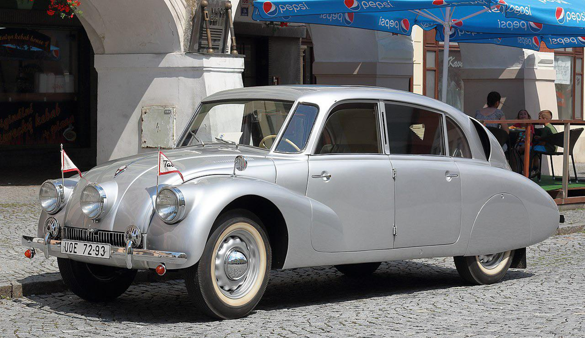 A vintage silver Tatra 87 car is parked on a cobblestone street in front of a building, with small flags on its fenders and blue Pepsi umbrellas in the background.