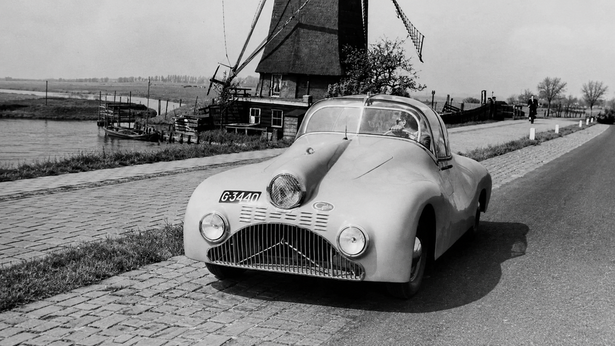 A vintage car with a unique, rounded design is parked on a cobblestone road next to a canal, with a windmill and trees visible in the background.