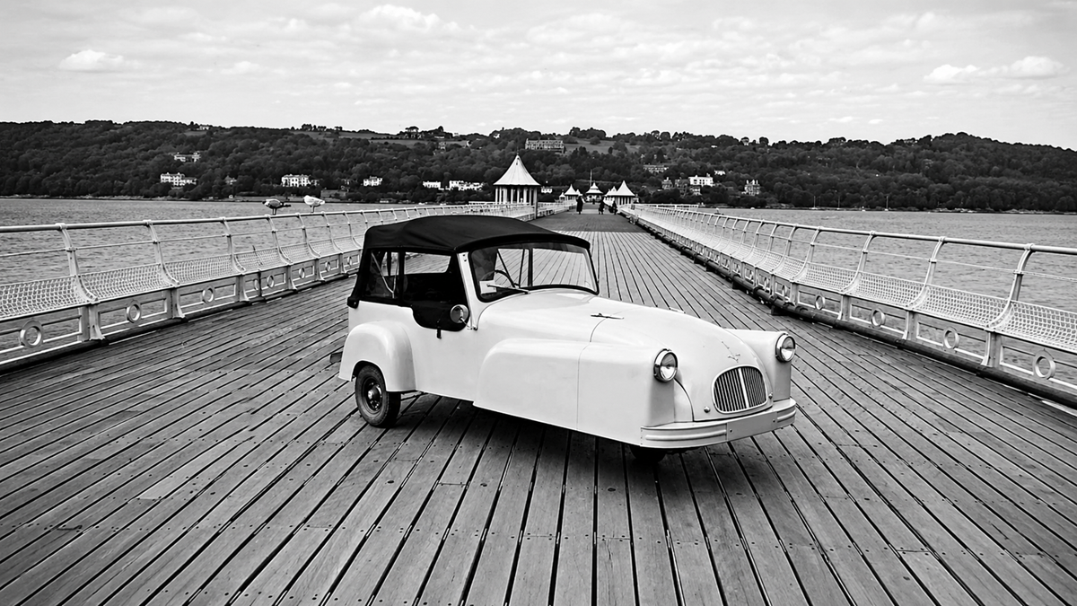 A small, vintage three-wheeled car with a black roof is parked on a wooden pier, with water and distant buildings visible in the background. The image is in black and white.
