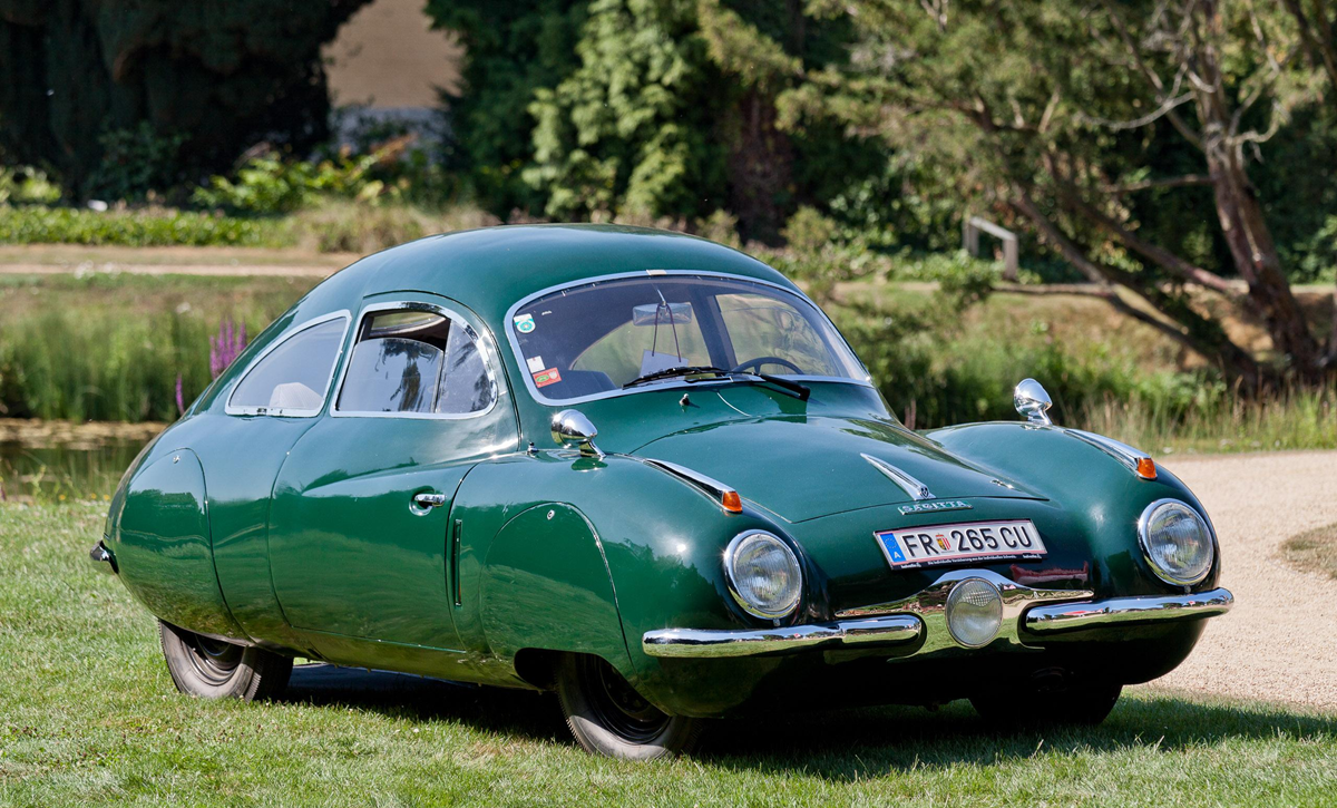 A vintage green car with a rounded, aerodynamic shape is parked on grass near a path, surrounded by trees and greenery.