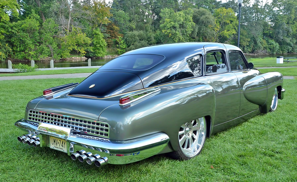 A shiny silver vintage car with a sleek, curved design and prominent chrome details is parked on green grass near a river and trees, under a clear sky.