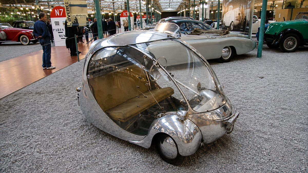 A small, bubble-shaped, silver car with transparent doors is displayed in a museum among other vintage cars. People are observing the exhibits in the background.