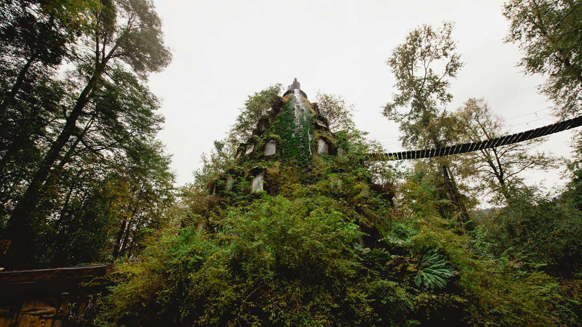 A moss-covered, cone-shaped building surrounded by dense forest, with a rope bridge extending from its upper level and greenery climbing up its walls under an overcast sky.