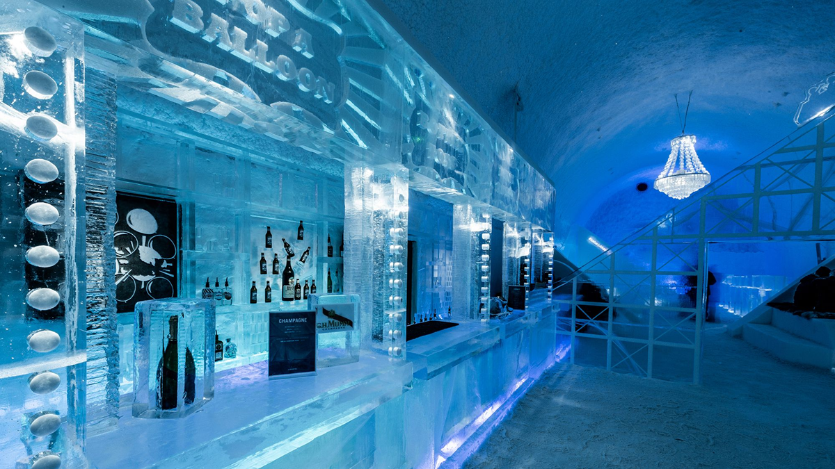 An ice bar with counters, shelves, and walls made of ice, illuminated by blue lights. Bottles and glasses are displayed on the ice shelves, with a chandelier hanging from the ceiling.