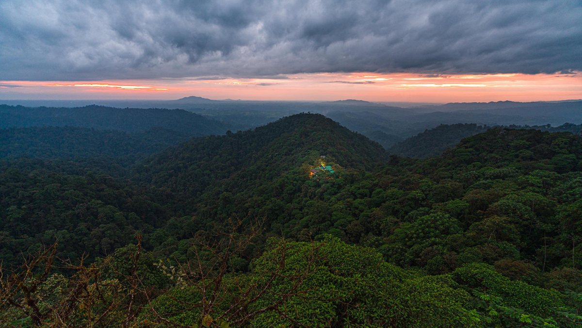 Dense green forested hills under a dramatic cloudy sky at sunset, with a cluster of lit buildings nestled among the trees in the center of the scene.