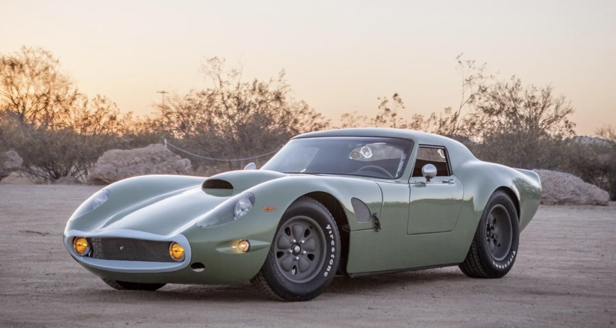 A vintage-style green sports car with rounded lines, hood scoop, and round headlights is parked on a sandy terrain at sunset, surrounded by desert plants and rocks.