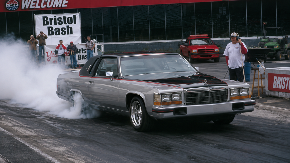 A custom Cadillac burns rubber at a drag strip, producing thick tire smoke, while a person with a headset stands nearby. A “Bristol Bash” sign is visible in the background.
