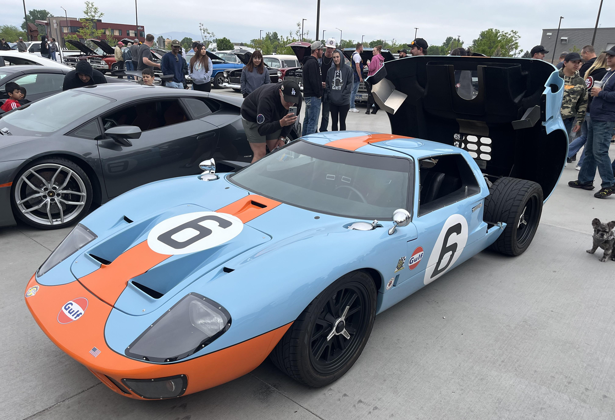 A blue-and-orange vintage race car with the number 6 is parked at a car show. People gather around, some looking at the engine, with a gray sports car and a small dog nearby.