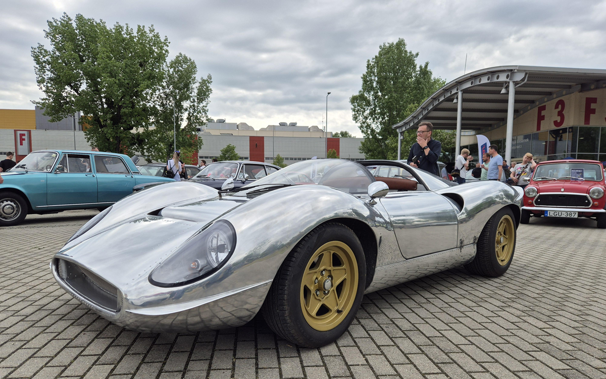 A shiny silver vintage sports car with gold wheels is parked at a car show. Several other classic cars and people are visible in the background under cloudy skies.