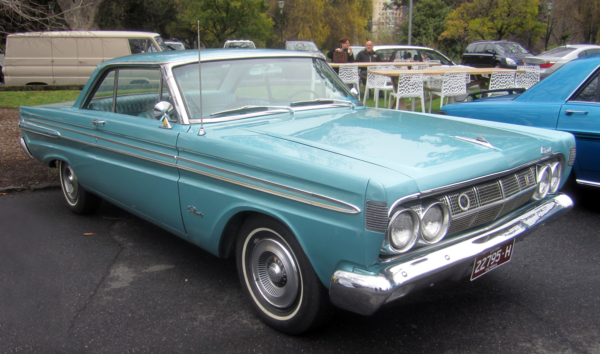 A vintage light blue classic car with chrome details is parked outdoors. There are white chairs and tables in the background, along with another blue vehicle and trees.