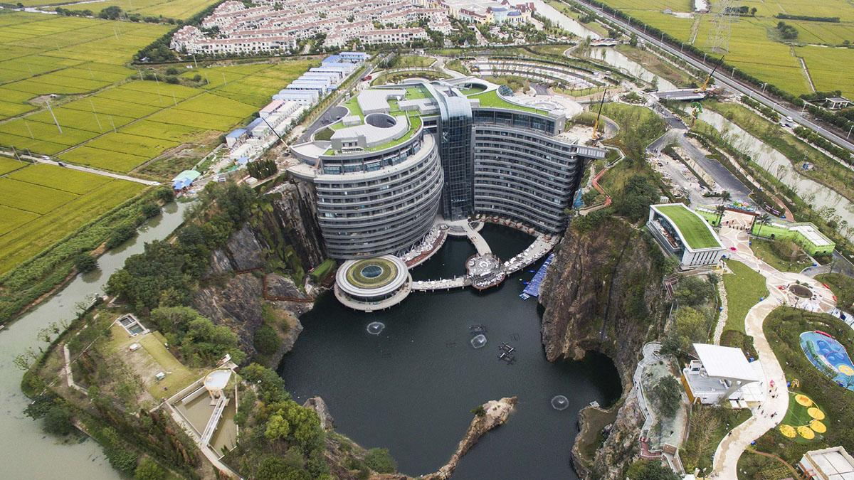 Aerial view of a large, modern hotel built into the side of a quarry with water at its base, surrounded by greenery, roads, and nearby buildings.