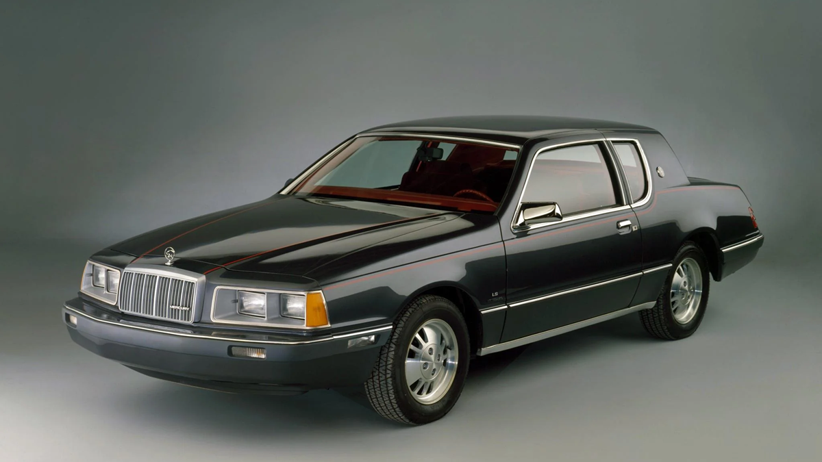 A dark-colored, two-door vintage car with chrome trim and a boxy design, photographed in a studio with a plain gray background.