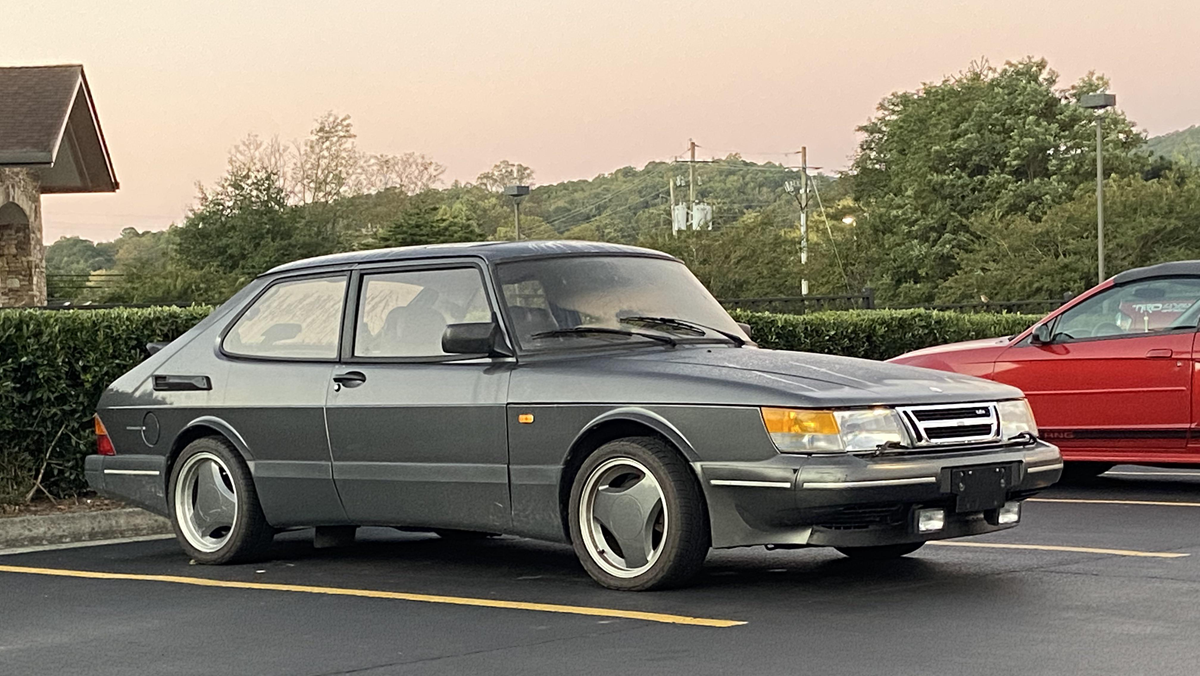 A gray vintage Saab 900 two-door car is parked in a lot, with trees and hills in the background, next to a red vehicle.