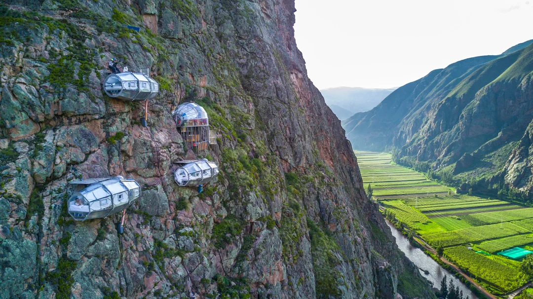Three transparent pod-like hotel rooms are attached to a steep mountainside, overlooking a lush green valley and river far below.
