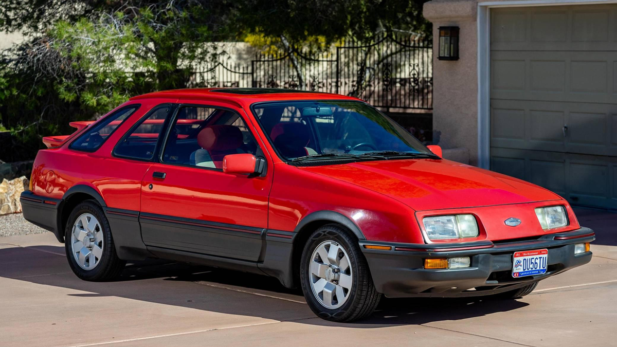 A bright red vintage coupe with black trim and silver wheels is parked in a driveway near a garage and wrought iron gate. The car has pop-up headlights and a blue and white license plate.