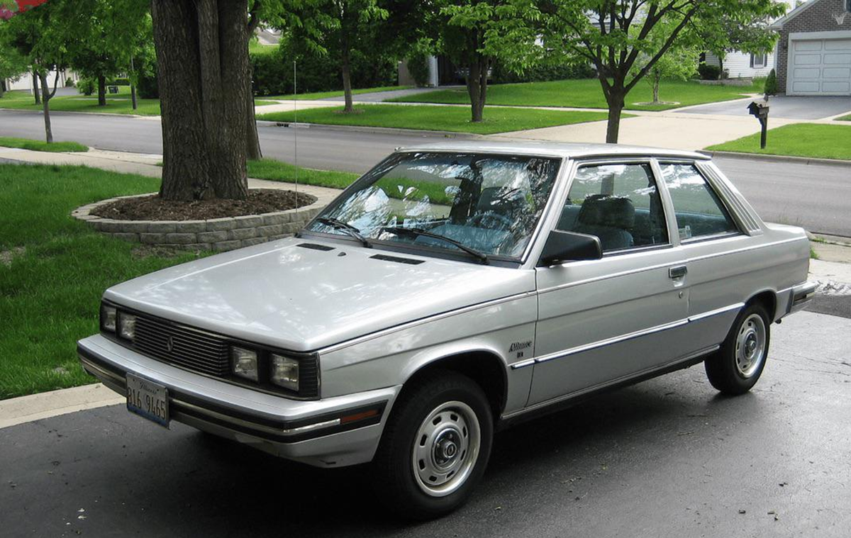 A silver two-door car from the 1980s is parked in a driveway near green grass, trees, and suburban houses on a cloudy day.