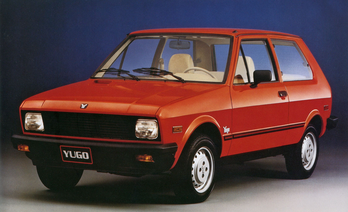 A red Yugo hatchback car is parked on a plain surface with a blue background. The car is viewed from the front left side, showing its boxy shape, black grille, and “YUGO” license plate.