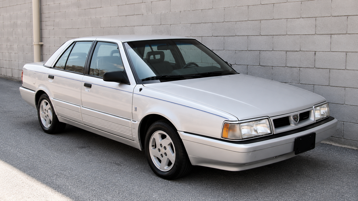 A silver four-door sedan is parked next to a gray concrete block wall on a sunny day. The car appears to be from the late 1980s or early 1990s.