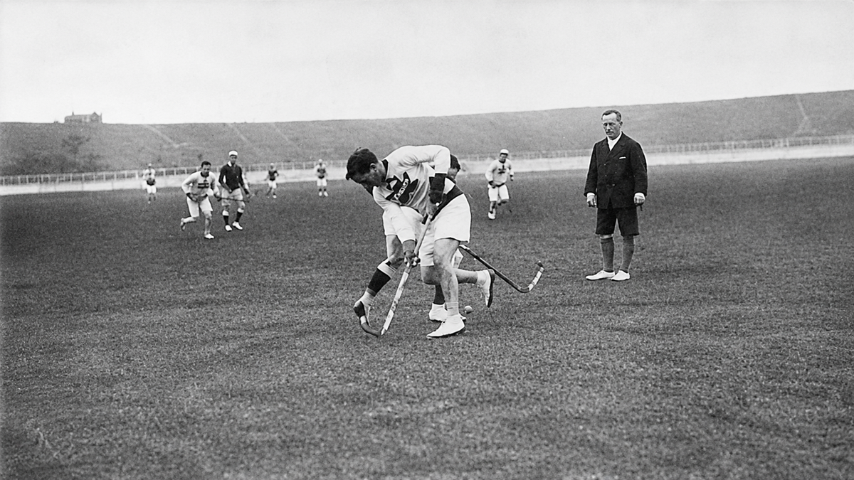 Two women compete for the ball in a field hockey match as other players and a referee watch. The scene takes place on a large, grassy field under an overcast sky.