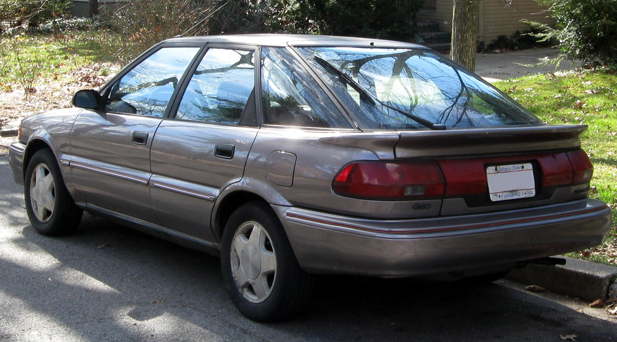 A brown four-door sedan is parked on the street next to a grassy area with trees and fallen leaves. The car appears to be an older model, in decent condition.
