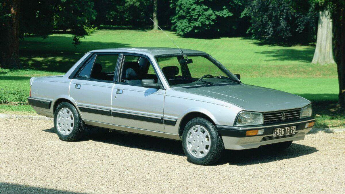 A silver, four-door sedan with black trim parked on a gravel path, surrounded by green grass and trees on a sunny day.